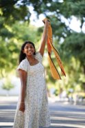 A young indian woman wears a long white dress with small floral details and a white cord around her neck. She smiles and proudly holds up a gold UC Davis graduation stole. 