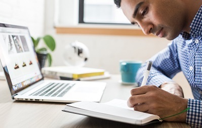 person sitting at a computer writing notes in notebook
