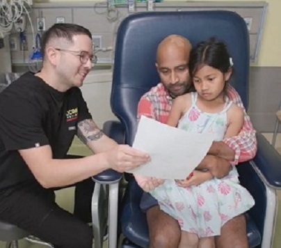 Father and daughter sit on seat while man in black shirt shows them a paper