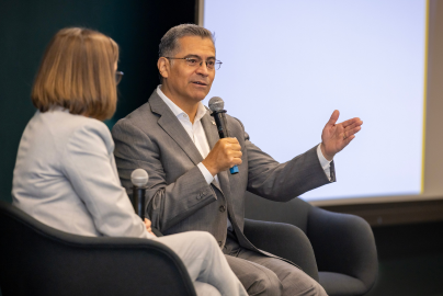 Courtney Lyles and Xavier Becerra are seated on a stage while hoding microphones in a conversation.