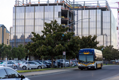 A bus drives on a road next to a glass building and parking lot. 