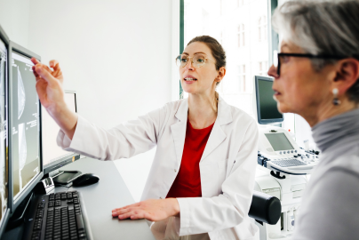 Two woman look at computer screens while one wearing a medical coat points to the screen.