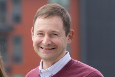 A smiling person wears a maroon sweater over a light blue collared shirt in front of an urban setting.