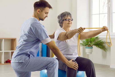 A male healthcare worker in scrubs kneels next to an older woman sitting on an exercise ball and stretching stretch bands with her arms.