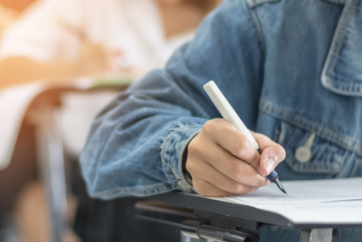 A student's hand is seen writing on paper at a desk surrounded by classmates.