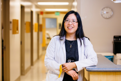 Elisa Tong smiles in a hospital hallway while wearing a white medical coat.