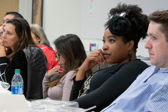 A group of people sit at a long table listening closely to a speaker off camera.
