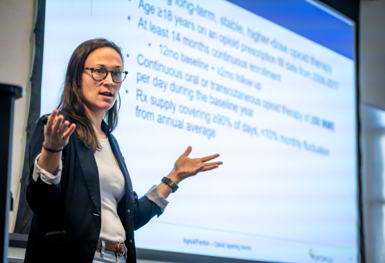 A woman in a black jacket and jeans speaks in front of a screen showing a presentation slide.