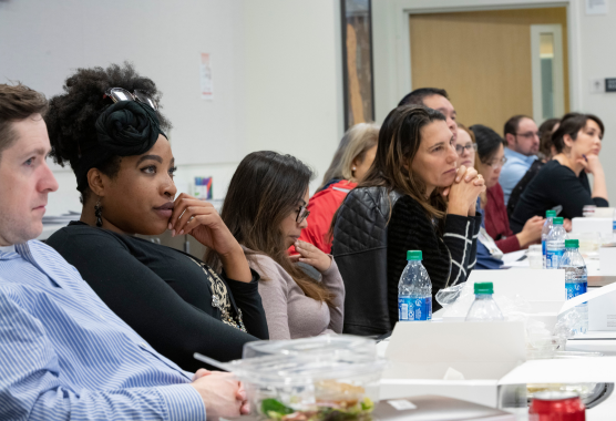A line of people seated at a long table with lunch wrappers and water bottles listens to a speaker off camera..