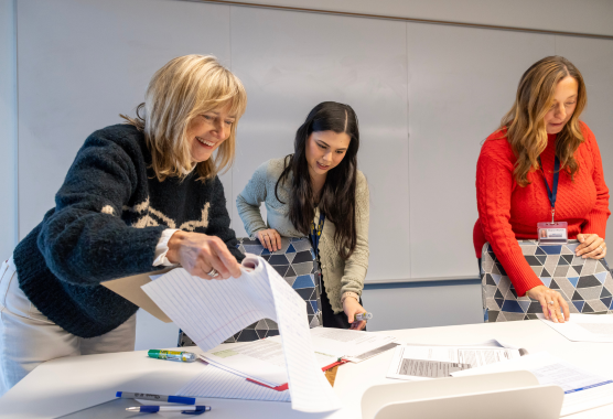 Three women stand around a table covered in papers. 