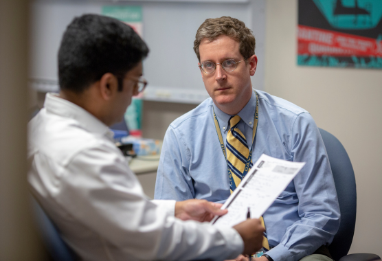 Stephen Henry is seen sitting in a chair listening to a man with his back to the camera.