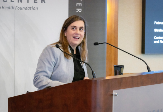 A person in a grey coat over a black shirt speaks behind a podium with a microphone attached..
