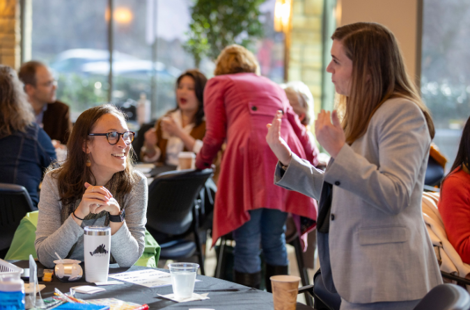 Two women talk with one seated at a table and the other standing