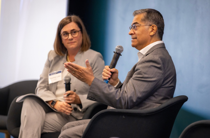 A man and woman wearing grey suits sit while holding microphones.
