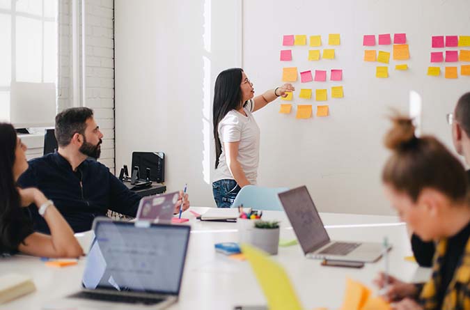 Group of people in a meeting room collaborating on a project, with one person placing sticky notes on a wall while others observe and take notes on la