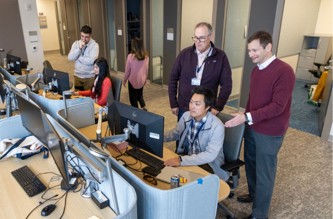 A group of people working at computers in an office setting. Two individuals are standing and observing someone seated at a computer, while another pe