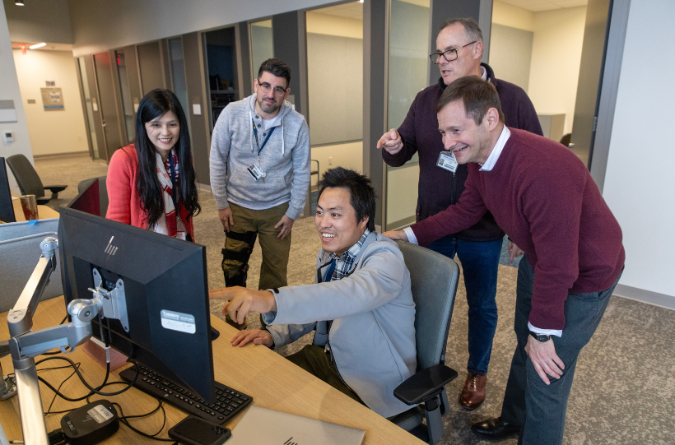 A man points to a computer screen with co-workers gathered around him. 