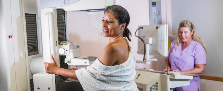 A Black woman in a steps up to a mammogram machine as a woman doctor stands nearby.