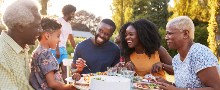 A multi-generational family sits around a table outdoors eating and smiling.