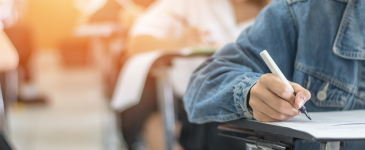 A hand with a pen is shown writing on a desk in a classroom full of students.