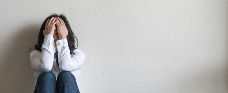 A woman sits on the floor against a wall with hands covering her face.