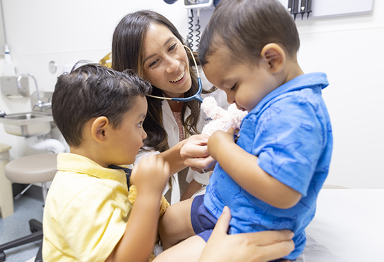 Female health care provider listening to young boy’s heart with older brother watching