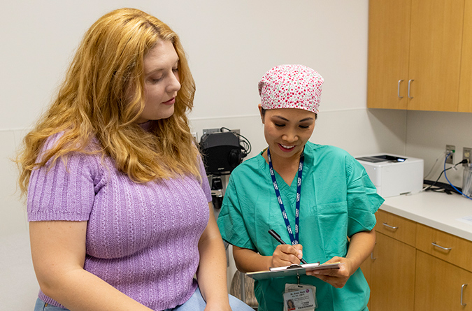 Woman sitting on exam table alongside nurse who is standing and explaining paperwork to her.