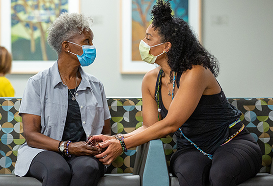 A masked young woman holding the hands of an older woman in a hospital waiting room.