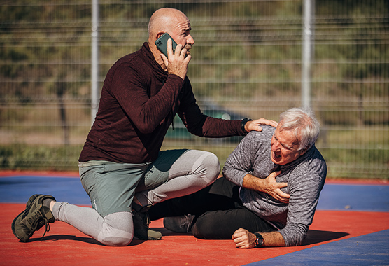 Man on the phone while another man lays on the ground grasping his chest.