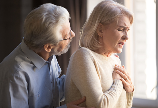 Older woman holding her chest trying to breath while older man is behind her checking on her.