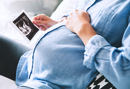 Pregnant woman keeping hand on belly and holding ultrasound image at home.