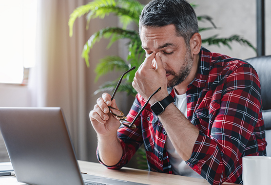 Man with his hand to his face with a headache as he sits at a desk.