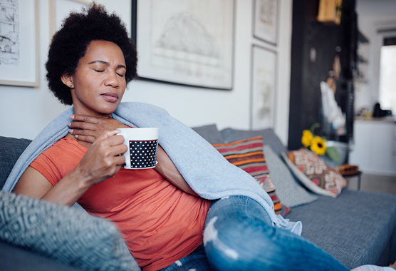 A sick woman drinking a hot beverage on a couch and rubbing her sore throat.