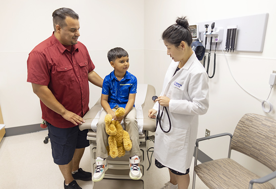 Young boy with leukodystrophy sitting next to his dad talking to a health care provider in a clinic room.
