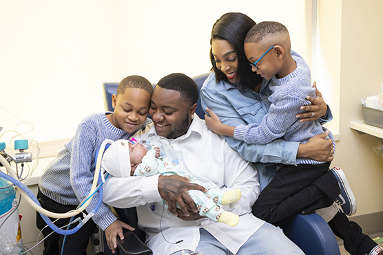 Dad holding NICU baby with mom and two older brothers gathered around smiling