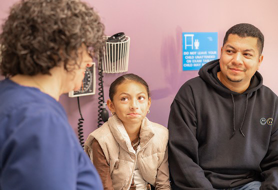 Young girl sitting on an exam room table next to her dad while talking to a nurse 