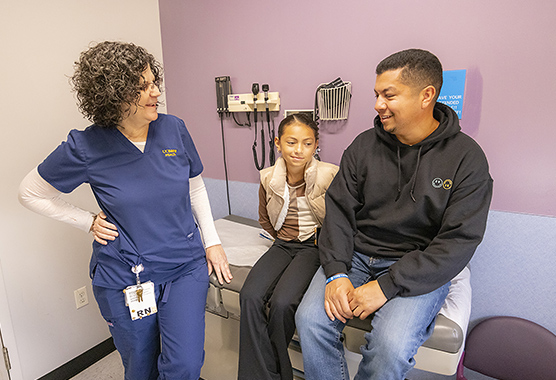 Girl and her dad sitting on an exam table talking to a female nurse.