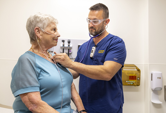 Older woman sitting on exam table with health care provider listening to her chest with a stethoscope.