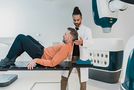 Man in an orange shirt being helped into place on a bed by a male medical provider