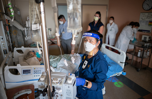 Nurse looking at infusion bag with patient behind her in hospital bed.