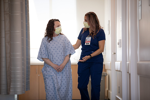 Female nurse and female patient wearing hospital gown walking down a hall