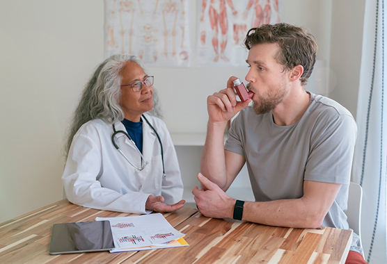 A man using an inhaler with a health care provider watching while sitting at a table.