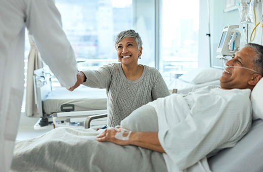 Woman shaking hands with surgeon while sitting next to man in hospital bed before minimally invasive surgery.