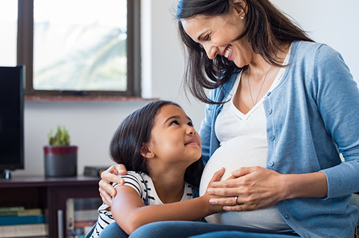 Pregnant mom smiling at young daughter who has her arms wrapped around mom’s belly 