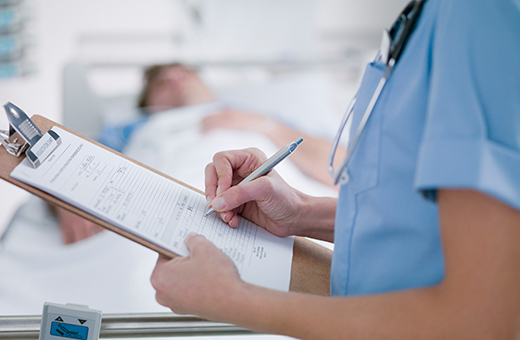 Patient in hospital bed after vascular surgery while health care provider writes notes using a clipboard.