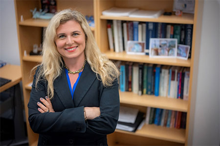 An individual with a blonde hair, smiling and crossing arms, wearing a dark jacket, standing in front of a bookcase.