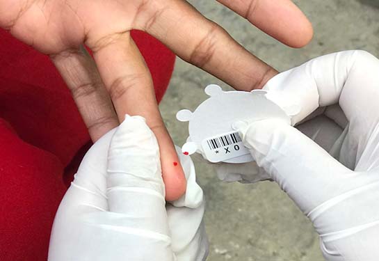 A pair of gloved hands is seen collecting a small amount of blood from a finger that has been pricked. 