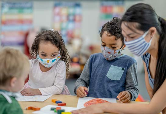 Children wearing masks sitting at a table with a teacher