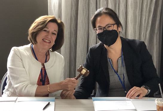 Two women sitting at a table share holding a brown wooden gavel. 