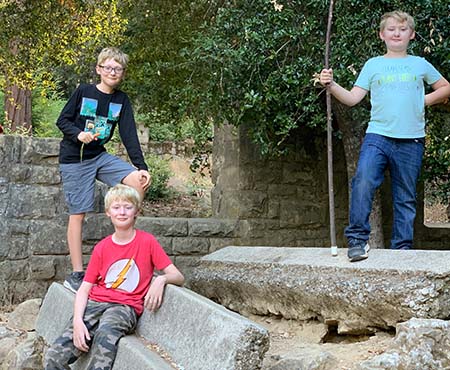 Two children stand on a small hill, while a third child sits toward the bottom of the hill. One child holds a stick in his hand like a walking staff. 